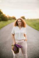 Young woman stands in white shirt on road with purple and pink lupins. Beautiful young woman with curly hair with bouquet of lupins. Sunset or sunrise, bright evening light photo