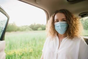 Stylish young carly woman in medical mask sitting on back seat of car on blurred background with sunset photo