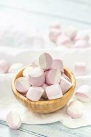 Portrait of a wooden bowl full of pink and white marshmallows with some scattered around on a white table cloth photo