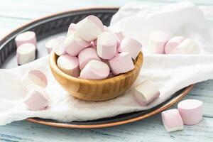 Wooden bowl full of pink and white marshmallows with some scattered around on a white table cloth, dark tray and white wooden table photo