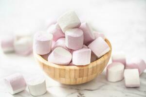 Wooden bowl full of marhsmallows with some scattered around on a marble surface in a light environment photo