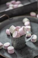 Portrait close up of a metallic vintage tray, surface and cup with pink and white marshmallows inside the cup and scattered around the tray as well as in the metallic box in the background photo