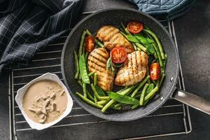 Top view of healthy cooked chicken and veg in a frying pan with a creamy mushroom sauce on the side, placed on a black background with a towel and a potholder photo