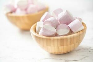 Pink and white marshmallows in a full wooden bowls placed on a marble surface with a focus on the bowl in the front photo