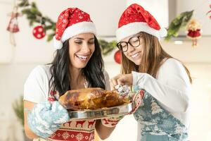 dos alegre cocineros, un madre y su Adolescente hija, en Navidad delantales con Papa Noel sombreros, horneado un ganso o pavo, el hija sabores eso foto