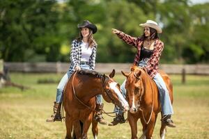 Two good-looking young women ride their horses, one pointing to the right while horses touch with lips photo