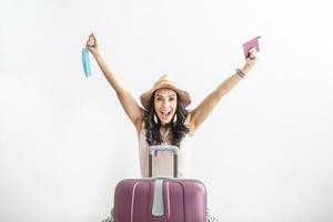 Happy female traveler stands in front of her suitcase, arms up as restrictions ease, holding passport in one and face mask in the other hand photo