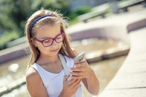 Young pre-teen blonde girl looks into the mobile phone in her hand sitting on the edge of a fountain on a sunny summer day photo