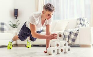 Fit young man creatively using excess toilet paper rolls for home plank and pushups workout in the living room. photo