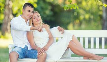 joven Pareja vestido en ligero ropa sentado en un blanco banco en un parque mirando el mismo dirección. chico señalando con un dedo, mientras el Pareja es sonriente foto