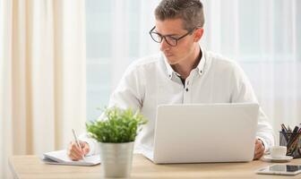 Male writes in his notepad with a notebook opened on his desk in front of him photo