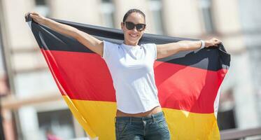 hermosa sonriente hembra ventilador en Gafas de sol sostiene un alemán bandera detrás su al aire libre foto
