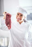 Female chef in professional white clothing holds a big chunk of red meat in her hand, smiling photo
