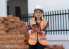 Civil engineer woman wearing hard hat and safety vest orange sittitng on red brick at the construction site,holding tablet using pencil write report, checking list buiding project. photo