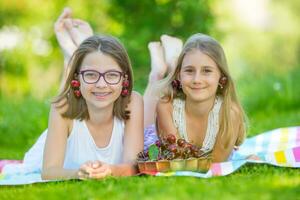 Two cute sisters or friends in a picnic garden lie on a deck and eat freshly picked cherries. photo