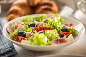 Healthy salad in a bowl with olives, raddishes, tomato, cheese and lean ham with croissants in the background photo