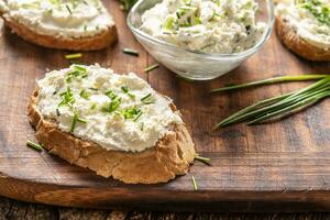 Slices of crusty bread and a glass bowl with a cream cheese spread and cut chives on a vintage wooden cutting board photo