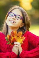 contento otoño niña sonriente y alegre participación otoño hojas. hermosa joven niña con arce hojas en rojo cárdigan foto