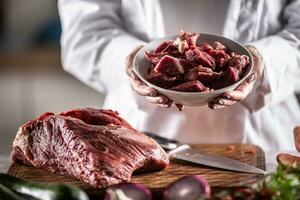 Small chunks of beef or pork for stew in a bowl next to a large piece of red meat on a cutting board photo