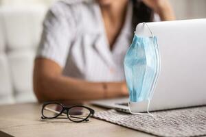 Detail of a face mask hung on a laptop and glasses put aside as woman keeps on working remotely after pandemic photo