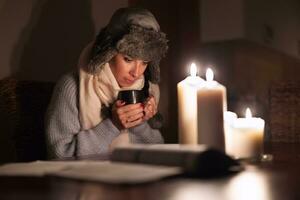 congelación joven mujer en invierno ropa calienta su manos en taza de té y luces con velas como energía apagones porque electricidad cortes foto