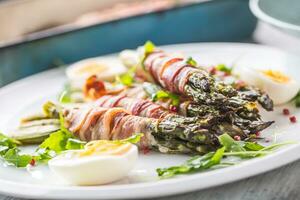 Fresh asparagus wrapped in bacon  on a white plate with arugula tomatoes and eggs photo