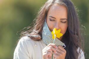 Pretty woman enjoys the smell of a flower with half of her face mask off, having closed eyes on a sunny day in the nature photo