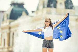 linda contento joven niña con el bandera de el europeo Unión en frente de un histórico edificio algun lado en Europa. foto
