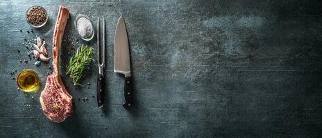 Raw tomahawk steak with fork knife salt spices garlic and herbs on a old rustic board - Top of view photo