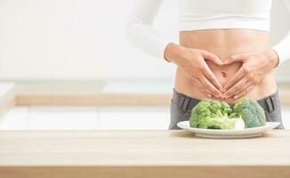 mujer con Deportes figura en su barriga muestra corazón forma. Fresco brócoli en plato en cocina mesa foto