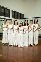 a group of Asian dancers stand side by side in white costumes in a room with a dance floor photo