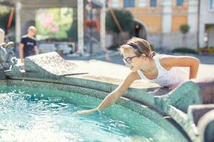 Young teenage girl splashes water from a fountain in the city on a hot summer day photo