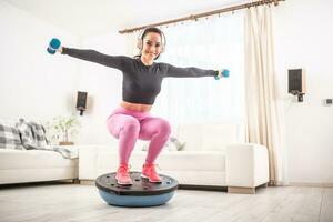 Home workout by a fit young female at home squatting on a balance ball with arms stretched to the side holding barbells photo