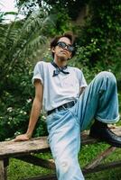 an Asian man in sunglasses and a white t-shirt sits on a wooden table in a park photo