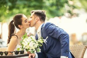 Joyful couple sitting and kissing each other after wedding. Bride holds elegant bouquet photo