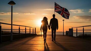 Pareja caminando debajo británico bandera en costero paseo en Inglaterra a puesta de sol. silueta concepto foto