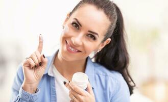 Woman holds face cream taken from a white packaging on her finger, smiling, cream also applied on her face photo