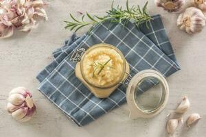 Top view aromatic garlic paste in a glass jar laid on rustic kitchen cloth with bulbs and peeled cloves and rosemary photo