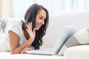 Smiling young good looking female with dark hair lying on a white couch with an open laptop, waving, having a video call photo