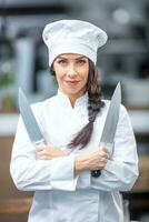 Good-looking female chef stands in the kitchen holding two knives smiling at the camera photo