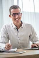 joven empresario vistiendo lentes sonrisas a el cámara mientras sentado a su oficina escritorio foto