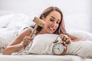 Female about to hit alarm clock with a hammer while lying in white sheets in her bed photo