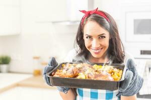 Beautiful wife holding a steaming hot tray in the kitchen, holding baked chicken and potatoes in her oven gloves, wearing apron and a red ribbon in her hair, smiling and looking into the camera photo