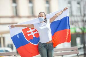 Smiling girl in a face mask holds a flag of Slovakia behind her on the street photo