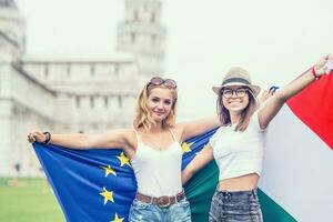 joven adolescente muchachas viajero con italiano y europeo Unión banderas antes de el histórico torre en pueblo Pisa - Italia foto