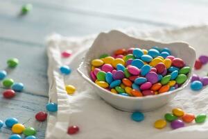 Smarties put in a white porcelain bowl and scattered around on a white clothe and a table photo