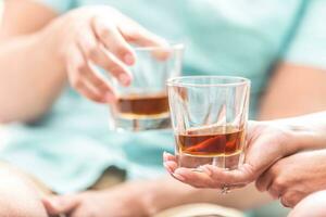 Man and woman hands toasting with glasses of whiskey brandy or rum indoors - closeup photo