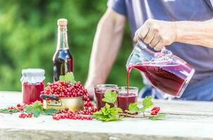 Man farmer in garden on table pouring currant syrup or wine. Red currants and products made of them on the table around photo