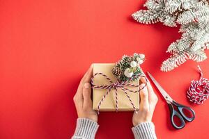 Navidad presente embalaje dentro un Respetuoso del medio ambiente papel con tijeras, enroscarse, y conífera árbol rama en un rojo antecedentes. foto