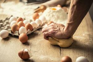 Hand of a man kneading dough by hand on a wooden surface with flour and eggs around photo